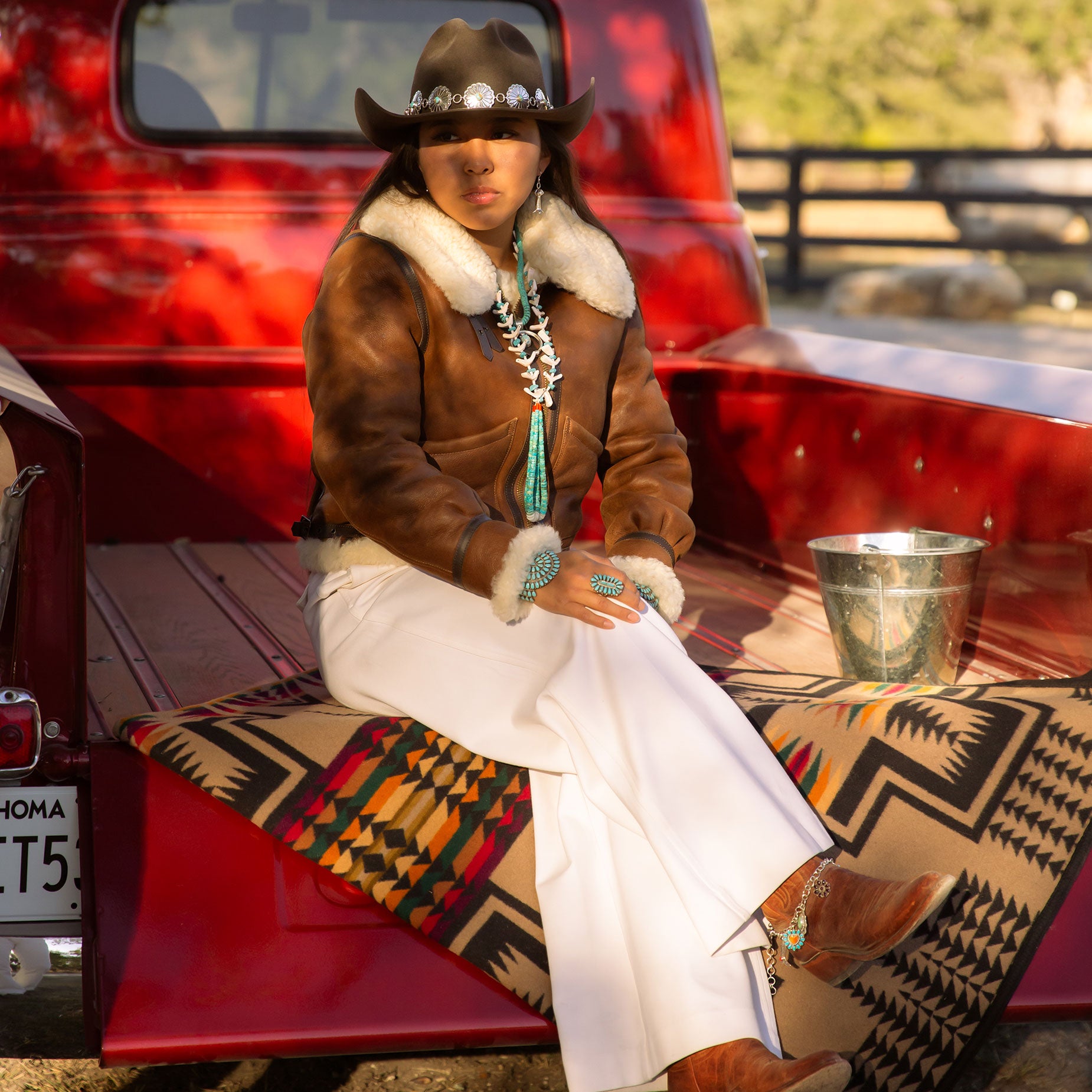 Girl in brown leather jacket, white pants, cowboy boots, and boot charms on the back of vintage red pickup truck.