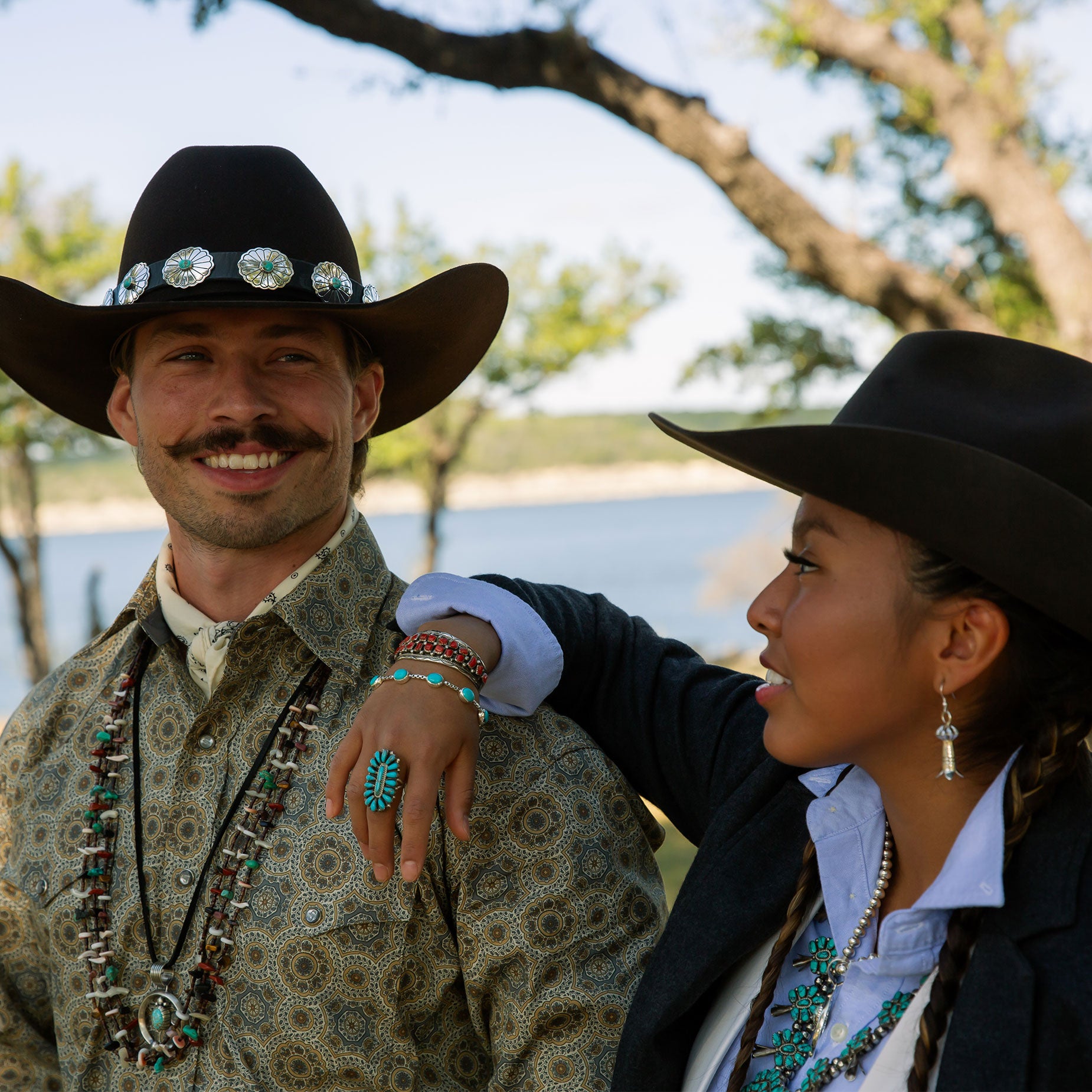 Two people wearing cowboy hats and turquoise jewelry in traditional attire outdoors with a scenic background.