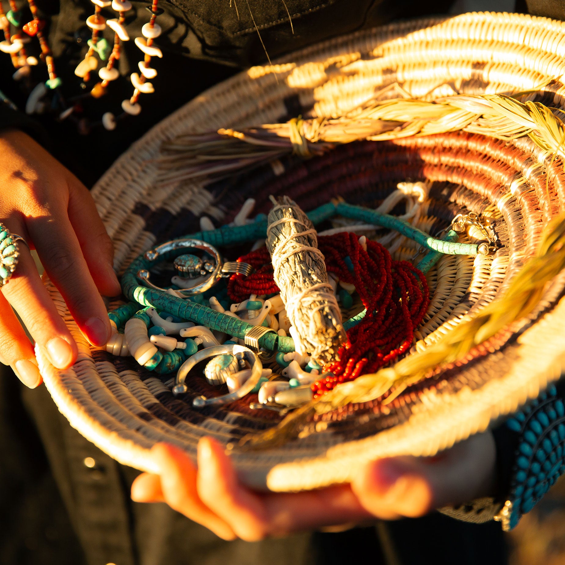 Person holding a navajo wedding basket with various items including navajo jewelry, heshi turquoise necklace, italian red coral, and a sage smudge stick.