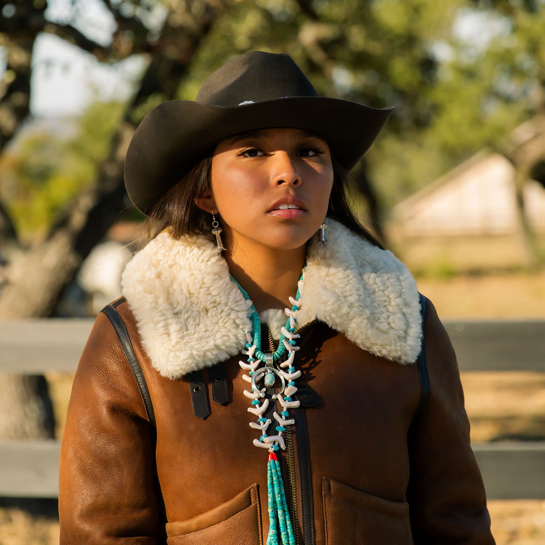 Person wearing a brown coat with a fur-lined hood and a cowboy hat outdoors on a ranch