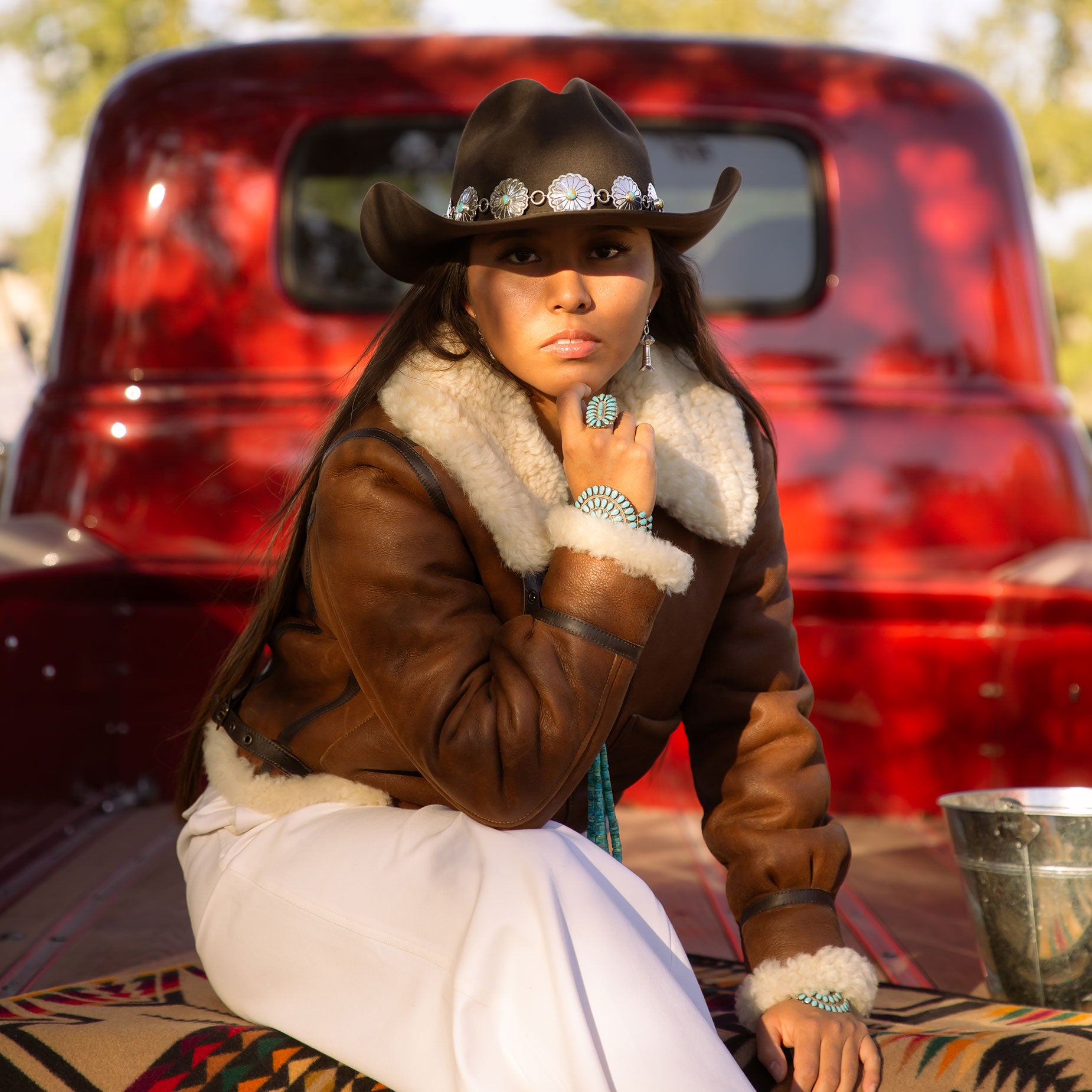 Person wearing a brown leather jacket with white fur lining and a cowboy hat, silver hat band, sitting on the tailgate of a vintage red truck.