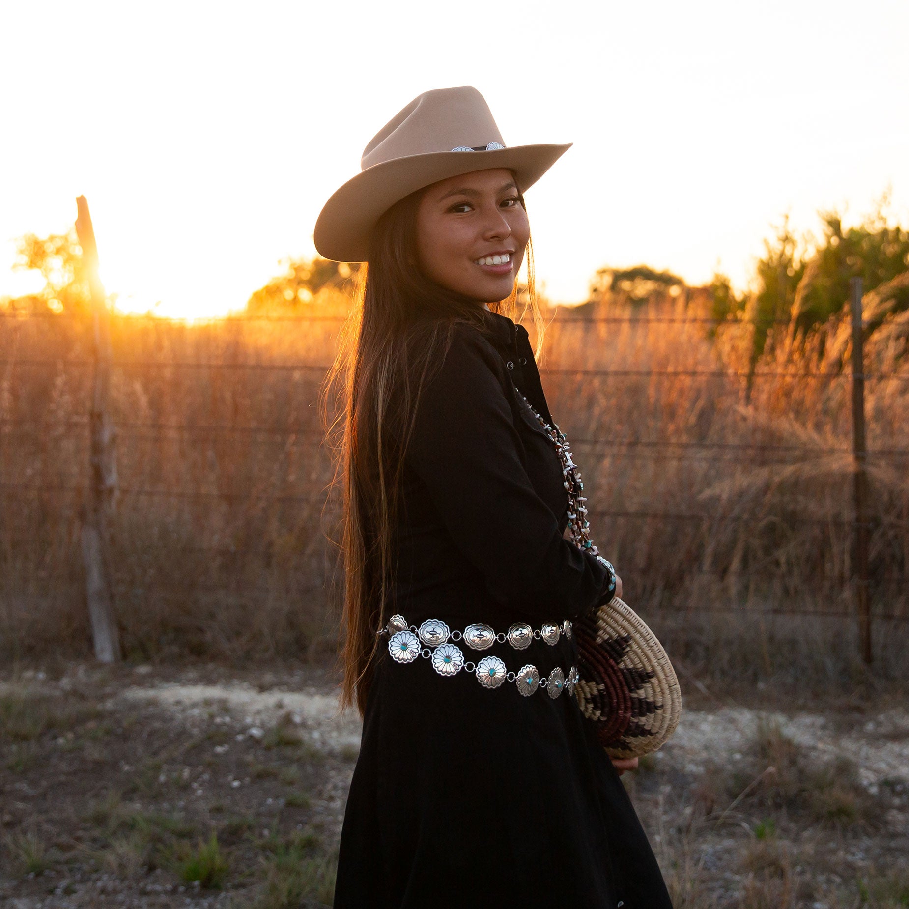 Woman in a stetson cowboy hat, and cowgirl & indians sterling silver concho belts, and black outfit with a navajo wedding basket standing in a field at sunset