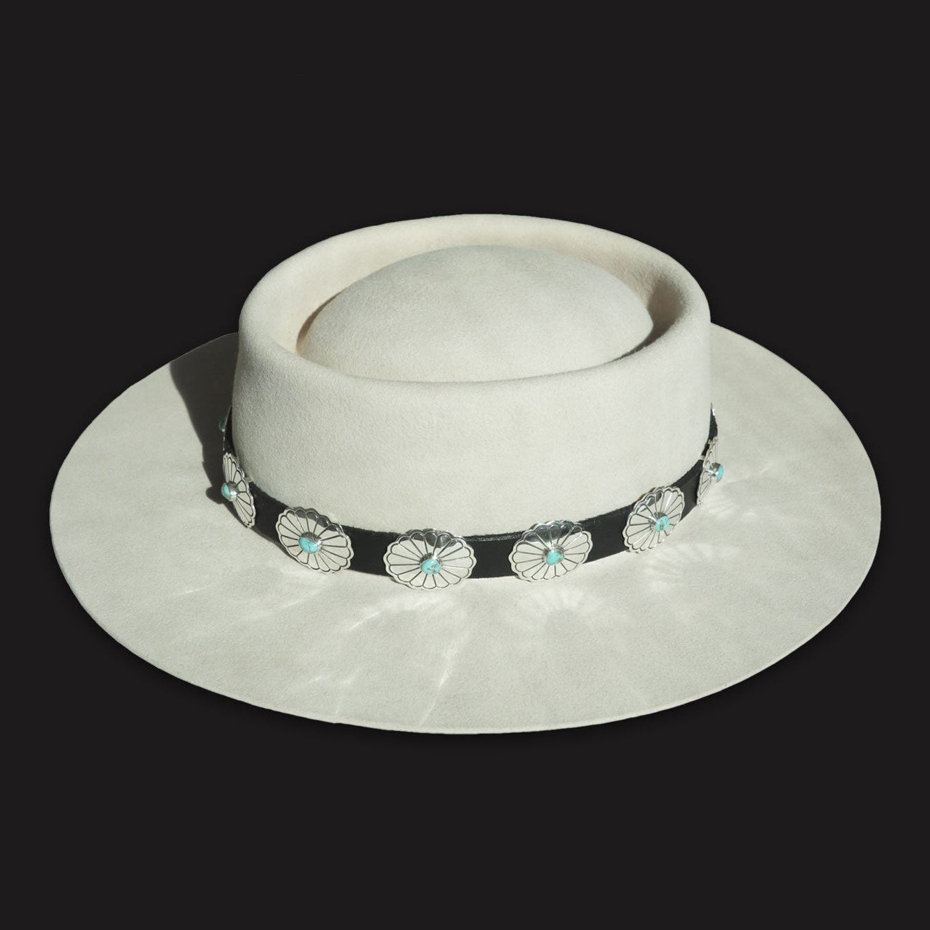 Rough Rider hatband displaying stamped silver conchos with turquoise on a dark leather band, shown on a light hat.