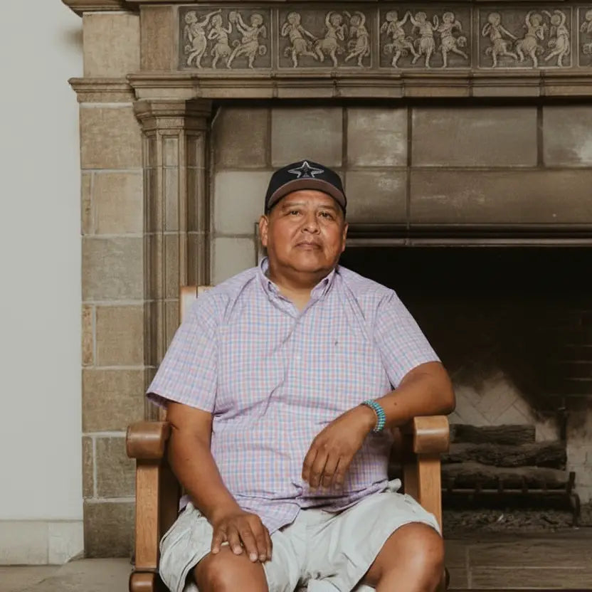 Navajo Silversmith, Lee Charley, with Dallas Cowboys Hat sitting in wooden chair in front of stone fireplace.