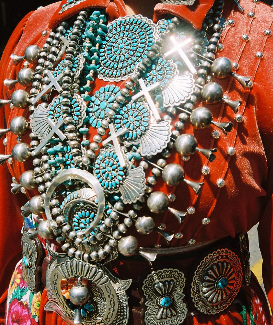 Layered authentic Native American jewelry featuring multiple sterling silver squash blossom necklaces with intricate turquoise cluster medallions and Naja Pendant, Navajo pearls (silver beads), silver crosses, and an award winning stamped concho belt buckle over traditional Navajo attire.