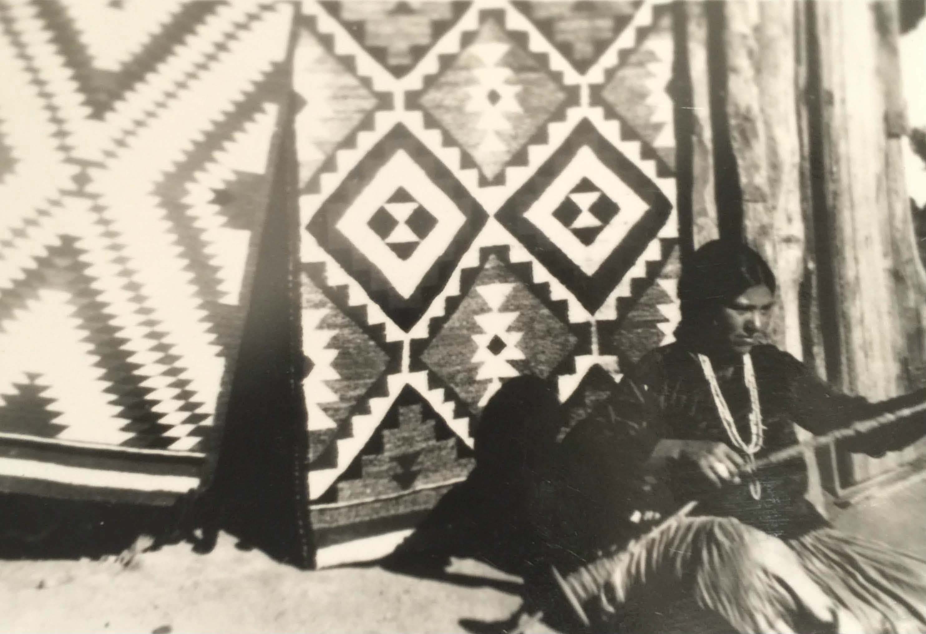 Navajo Rug Weaver, Tessie Skeet, sits beside her Navajo Rug on the dirt floor while demonstrating how to handspin wool using a spindle.
