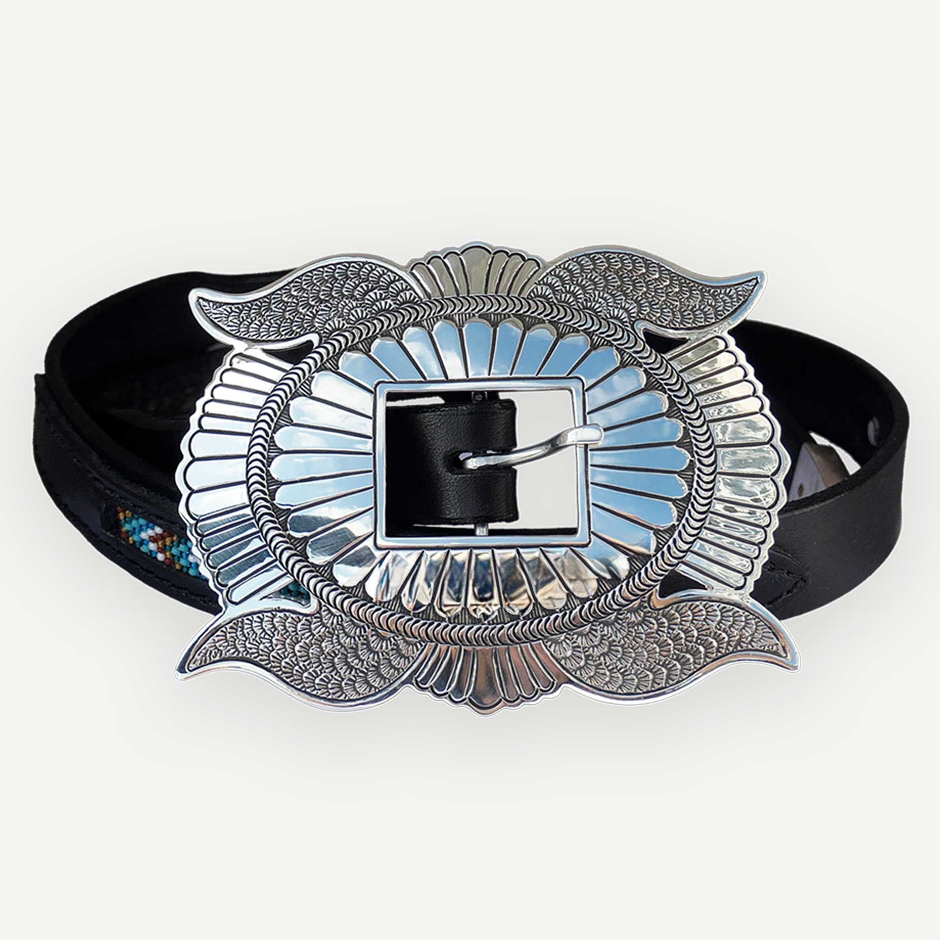A navajo concho belt buckle with a large ornate silver-tone buckle featuring wing-like flourishes and a blue inlay, paired with a black leather strap, shown on a light gray background.