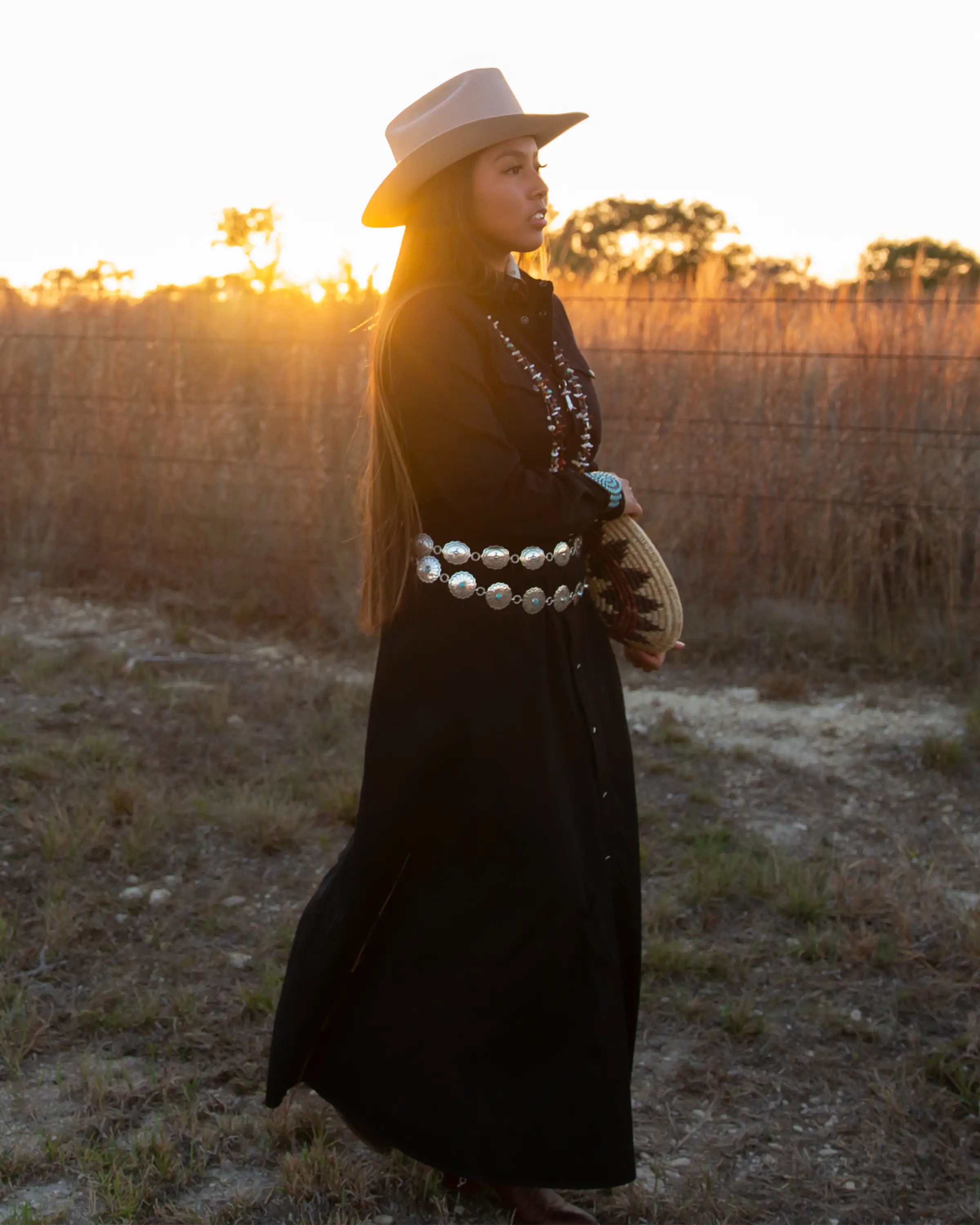 Woman in a black dress, concho belts, and cowboy hat, holding a navajo wedding basket, standing in a field with sunset in the background