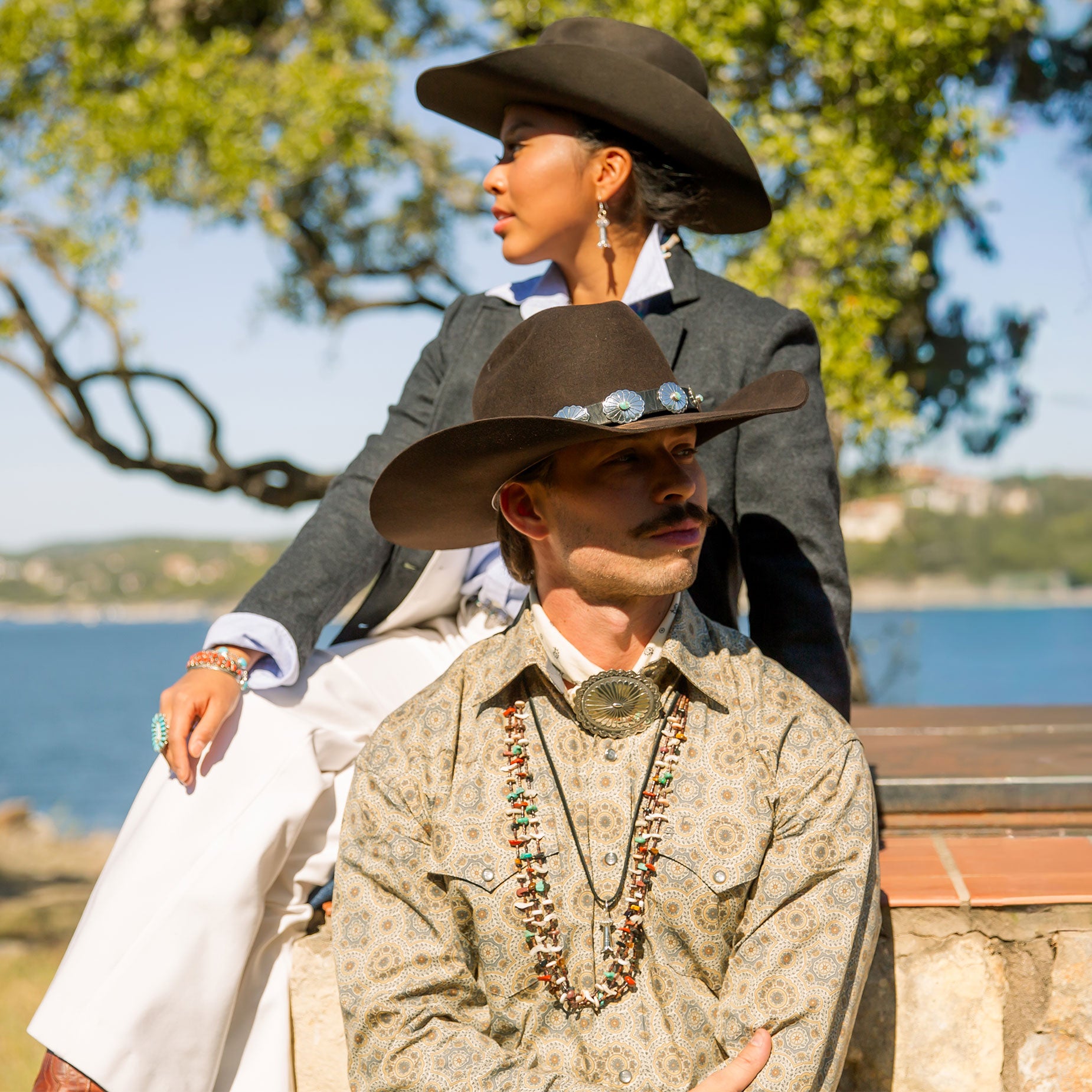Two people in cowboy attire sitting by a scenic lakeside.