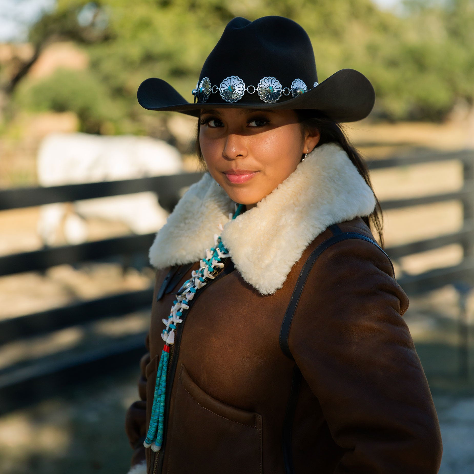 Girl wearing a cowboy hat and brown coat with a blurred background of trees and a fence.