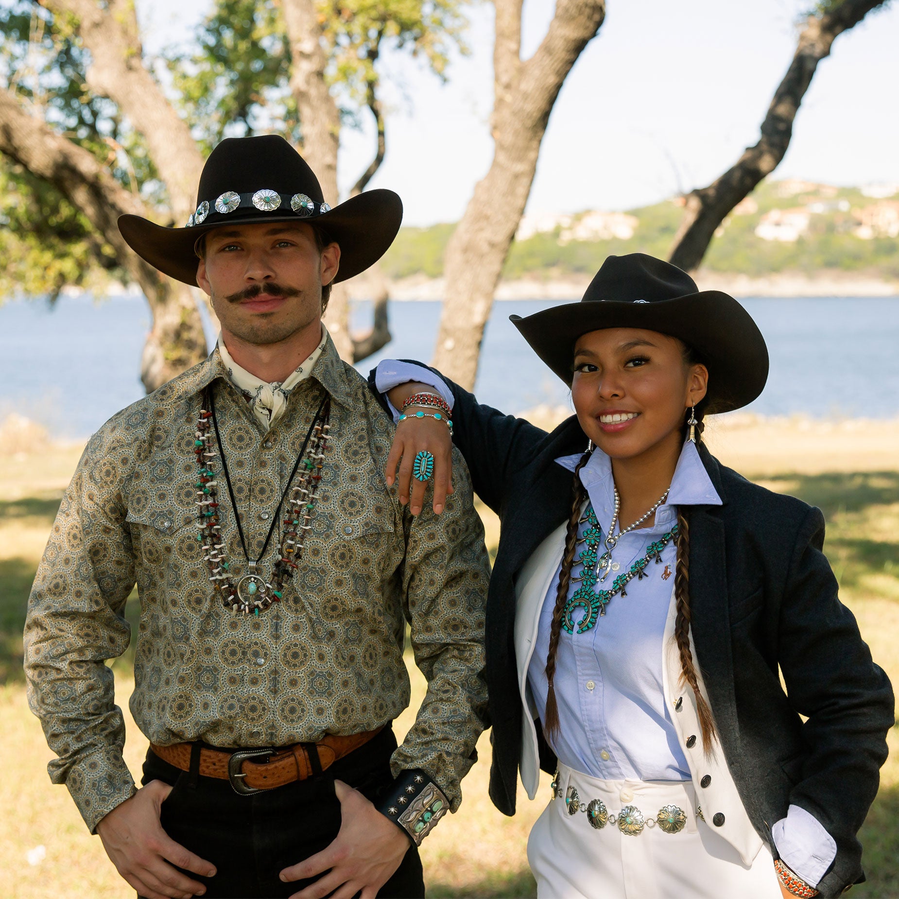 Two people in cowboy hats and western wear standing outdoors with a scenic background