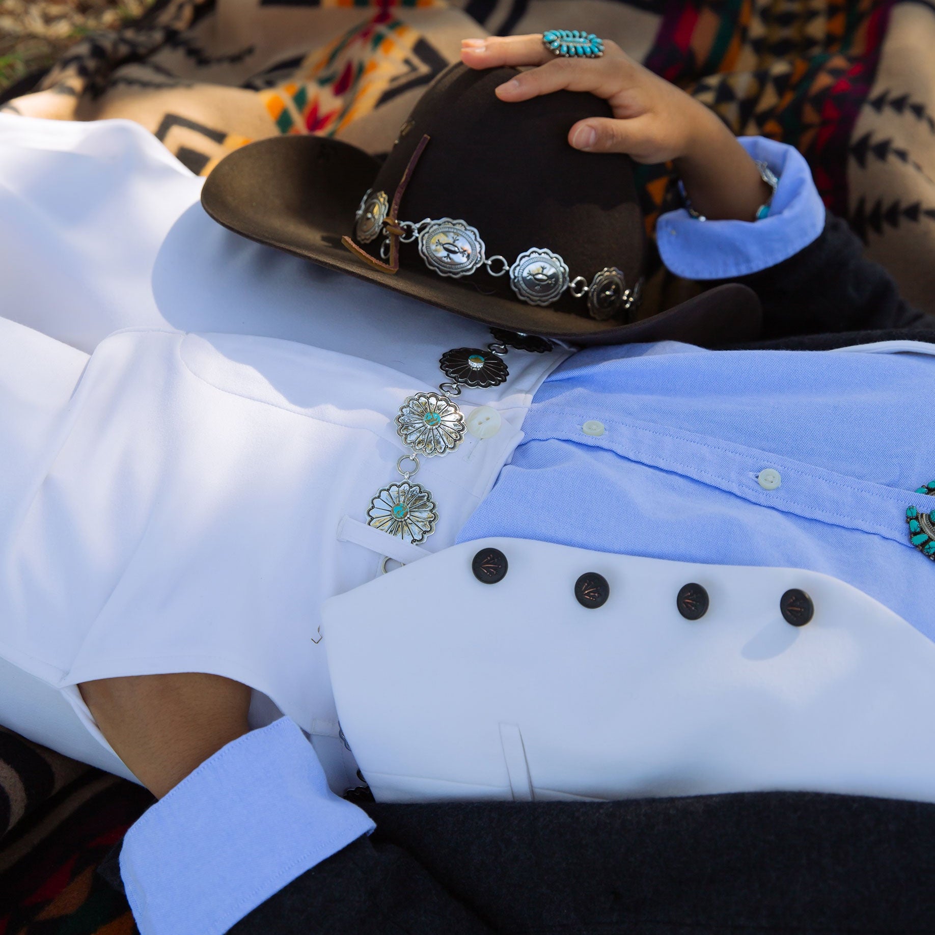 Person wearing a white vest with decorative buttons and a grey wool jacket, holding a brown cowboy hat with a navajo link hat band, and navajo concho belt in belt loops.