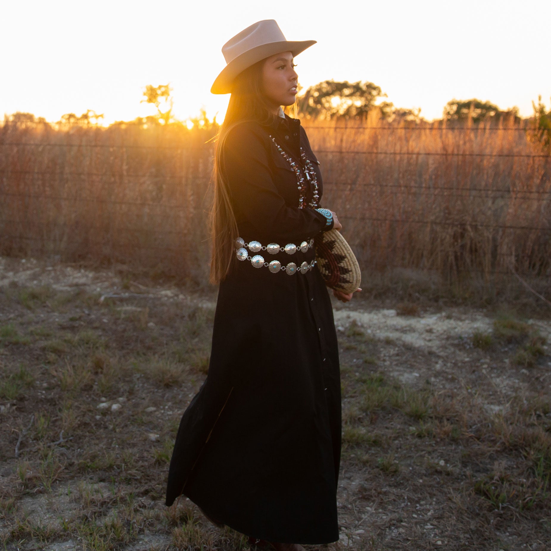 Woman in a long black dress, sterling silver concho belts, and hat standing in a field with sunset lighting.
