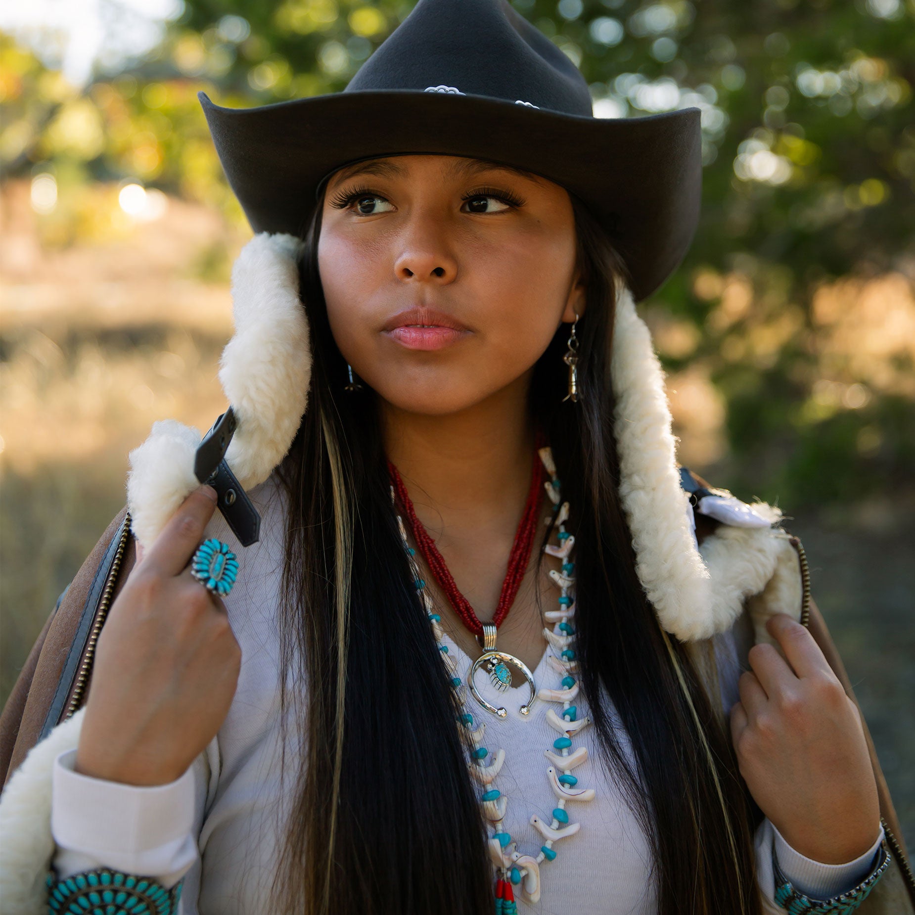 Woman wearing a black cowboy hat and fur-lined coat with a blurred natural background