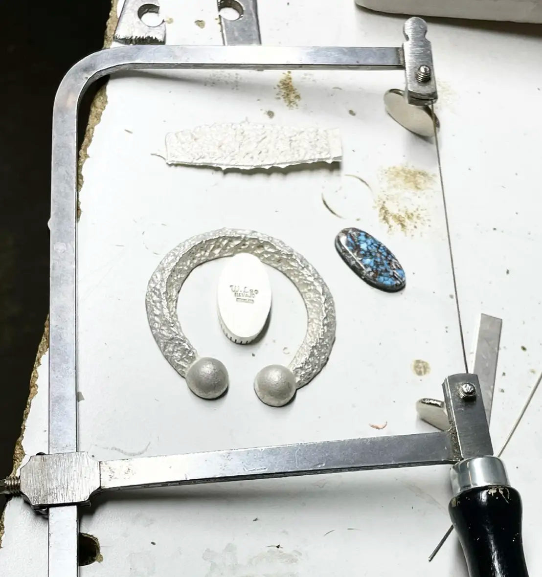 Jewelry-making process with silver naja pendant, turquoise gemstone, and tools on a white surface.