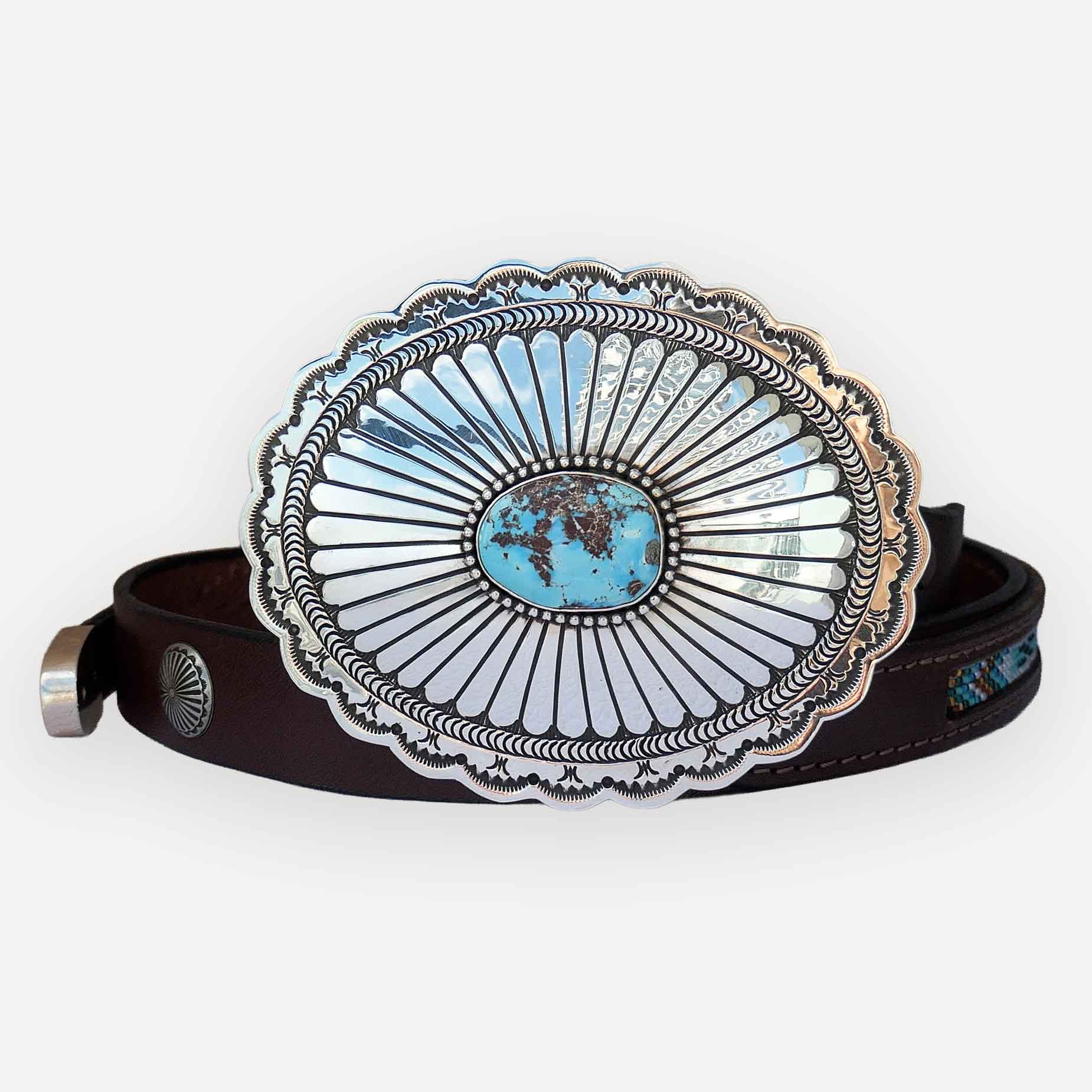 A western-style belt with a large oval silver-tone buckle featuring a turquoise center and sunburst detailing, attached to a dark brown leather strap.