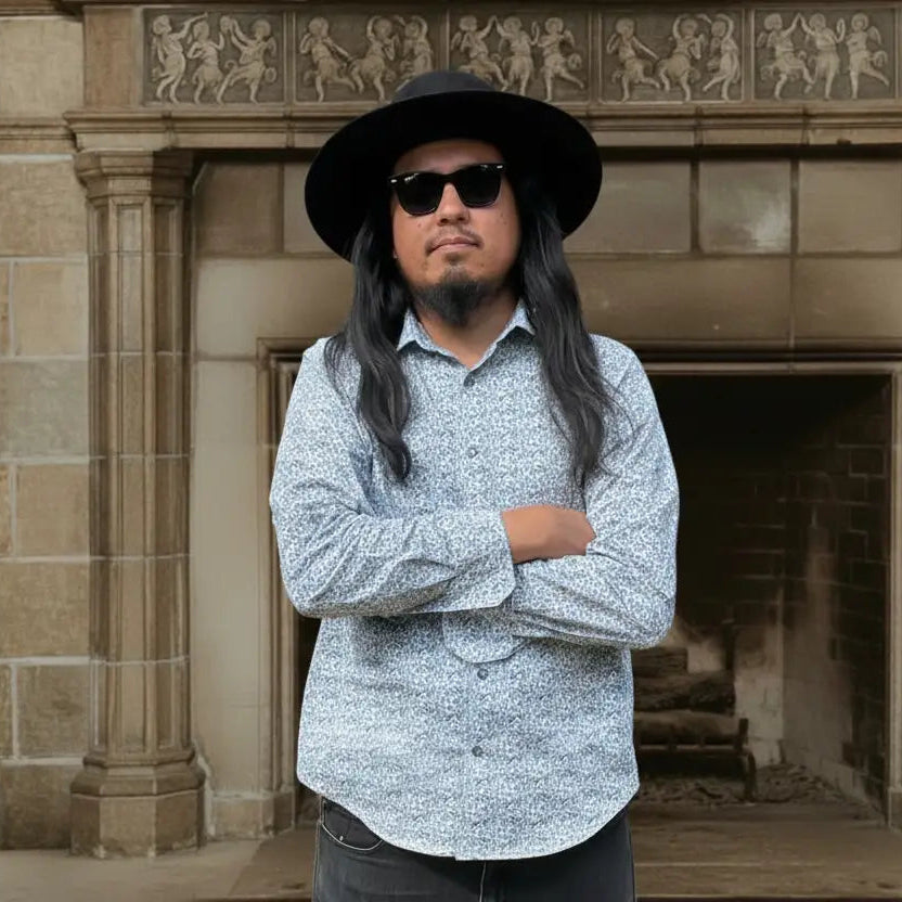 Diné Silversmith, Wyatt Lee Anderson, with long hair and beard wearing sunglasses and a hat, standing in front of a stone fireplace.